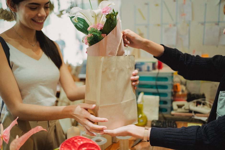 A florist at Hoxton Florist, dressed in a beige apron and white top, is handing over a bouquet of fresh white calla lilies and pink roses wrapped in pink tissue paper, inside a brown paper bag, to a c