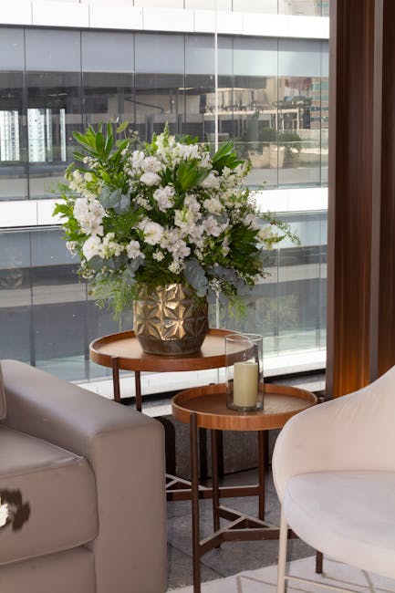 A large floral arrangement of white flowers, including roses and lilies, is displayed in a geometric metallic gold vase placed on a wooden side table in front of a window with a cityscape view. The bo