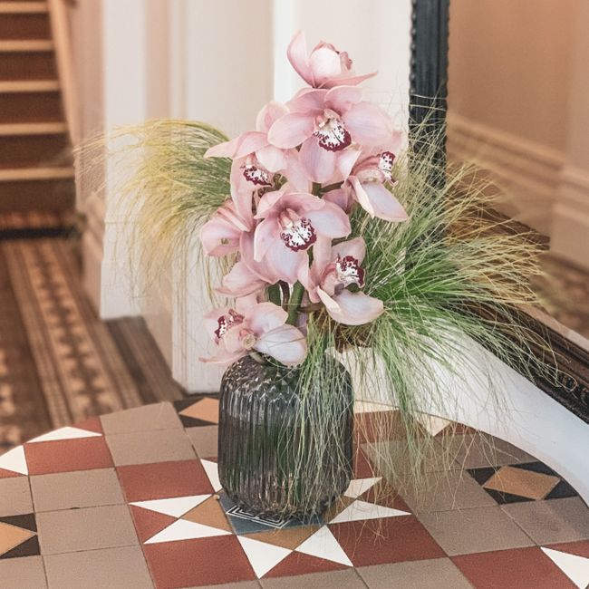 A tall floral arrangement featuring soft pink orchids with intricate, dark markings on their lips, placed in a dark, ribbed glass vase. The orchids are complemented by delicate, wispy green ornamental