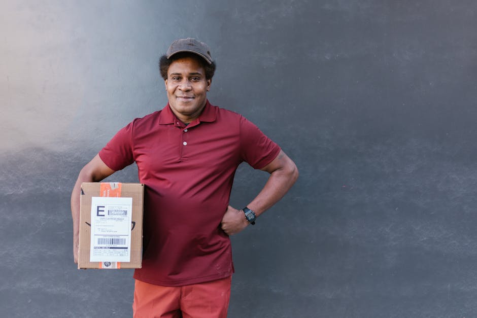 A male delivery person with medium brown skin and short curly hair, smiling, holding a brown cardboard parcel with a shipping label, standing outdoors against a plain, dark grey wall. He is wearing a 