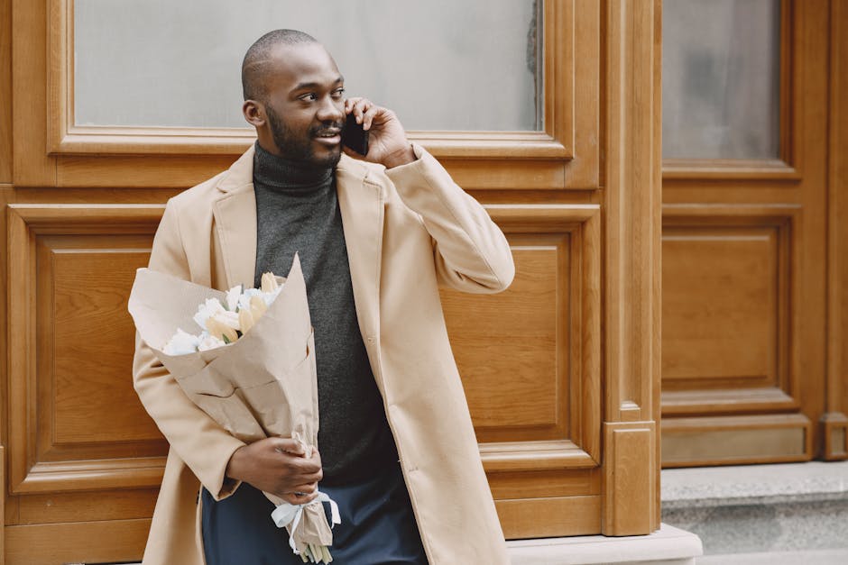 A smiling man with dark skin, wearing a beige overcoat and a dark turtleneck, is standing outdoors against a wooden-paneled wall. He is holding a bouquet of fresh white roses wrapped in brown kraft pa