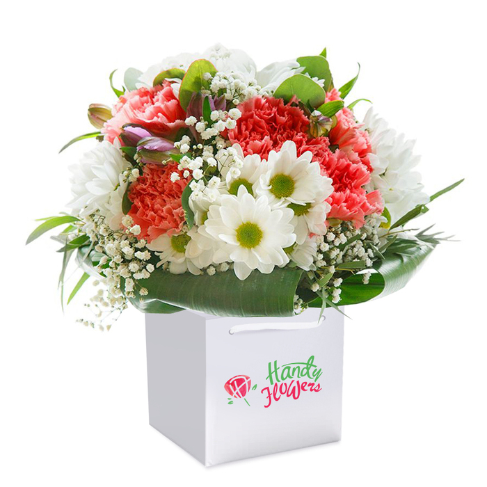 Set against a flawless white background, this lush and spirited bouquet rests gracefully in a square white gift bag, capturing the refreshing mood of a morning stroll through Hoxton's leafy streets. The bouquet itself features an exquisite balance: clusters of coral, peach, and vibrant red carnations, their ruffled petals brushing against clear, white daisy-like chrysanthemums with sparkling yellow-green centers. Tiny, cloud-like blooms of baby's breath (Gypsophila) are woven throughout, imparting a sense of softness and openness, while a few purple-tinged buds-perhaps preparing to bloom-offer just a whisper of depth and intrigue, like the unexpected charm of hidden spots along Kingsland Road. The arrangement is haloed by rich, dark green foliage and broad, glossy leaves, which gently curve around the bouquet's rounded shape and supply a natural, sophisticated border. The white gift bag, structured and sturdy with strong handles, suggests a ready-to-gift presentation-perfect for special occasions in Hoxton, whether birthdays, anniversaries, or heartfelt 'just because' gestures. The absence of distractions in the composition magnifies the freshness and vitality of the flowers themselves. This detailed florist-crafted bouquet becomes a visual invitation to pause and revel in Hoxton's unique blend of modern elegance and neighbourhood warmth, making it as fitting for a celebration delivered to an artist's loft as for a thoughtful surprise on a friend's doorstep nearby.