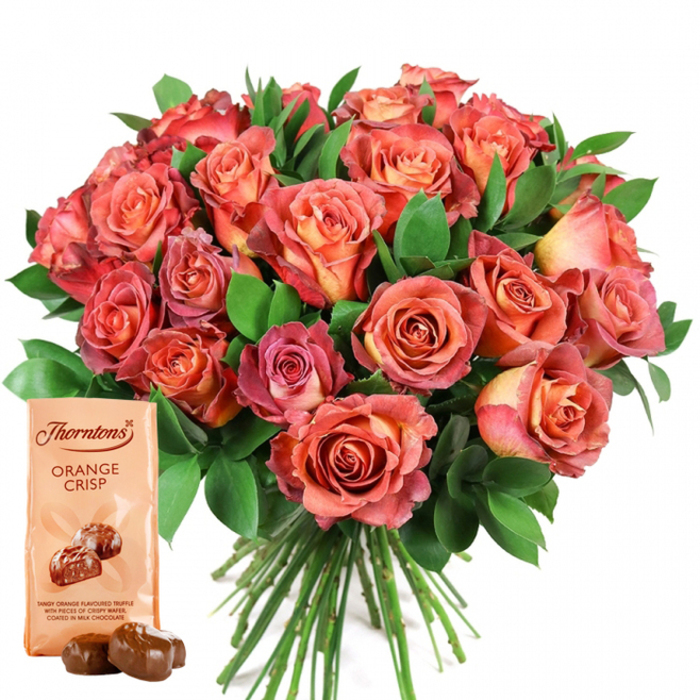 At the heart of this image is an opulent, rounded bouquet of fresh roses-artfully composed and bursting with warmth-crafted with care at a local Hoxton florist. The roses create a mesmerising gradient, their tightly furled centres glowing in soft peach and golden orange, opening out into petals edged with coral, rosy pink, and hints of deep, almost reddish hues. This bicolour and ombre effect lends a natural sophistication, while each bloom's velvety texture is subtly highlighted in the even, daylight-filtered setting. Interwoven between the radiant blooms, glossy ruscus or eucalyptus leaves add density and luxurious depth, their rich green tones perfectly framing the floral display. In the lower-left foreground, thoughtfully positioned for visual harmony, stands a Thorntons 'Orange Crisp' chocolate pouch-the cream-toned packaging echoing the bouquet's inner petals and prominently displaying the brand's logo. An inviting image of chocolate truffles and their tempting description-"tangy orange flavoured truffle with crispy wafer, coated in milk chocolate"-promise indulgence. Resting before the pouch, two real truffles glisten, moments away from savouring. The bouquet's green stems extend neatly below the full head, a testament to fresh, local floristry. This arrangement, set against total white, keeps attention on the vivid colours and delicious chocolates-a pairing perfect for special occasions or meaningful gestures, echoing the warmth and hospitality of Hoxton's lively community, from Columbia Road Flower Market to the vibrant shops on Kingsland Road.